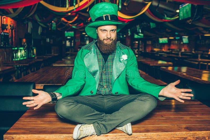 Nice handsome young bearded man in St. Patrick`s suit sit with legs crossed on table in pub. His costume green. Guy pose. royalty free stock images Nice handsome young bearded man in St. Patrick`s suit sit with legs crossed on table in pub. His costume green. Guy pose. royalty free stock images