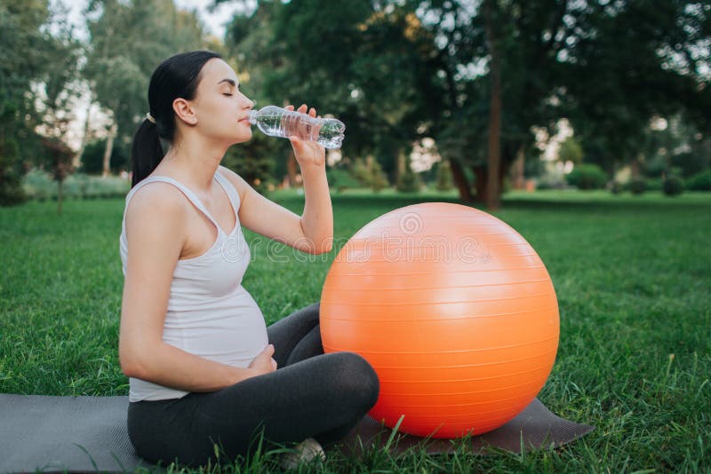 Nice young oregnant woman drinking water in park. She sit on yoga mate and hold one hand on belly. Orange fitness ball stock photo Nice young oregnant woman drinking water in park. She sit on yoga mate and hold one hand on belly. Orange fitness ball stock photo