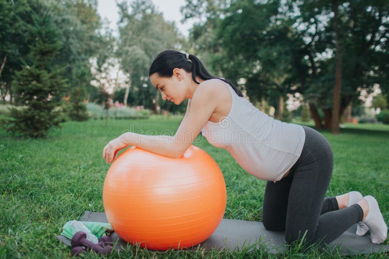 Nice young pregnant woman having workout in park. She stand on knees and look down. Model lean to big orange fitness royalty free stock photo Nice young pregnant woman having workout in park. She stand on knees and look down. Model lean to big orange fitness royalty free stock photo