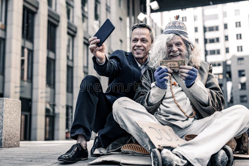 Excited old homeless. Vain rich men in costume making selfie of donation money to desperate poor man. Excited old homeless. Vain rich men in costume making selfie of donation money to desperate poor man