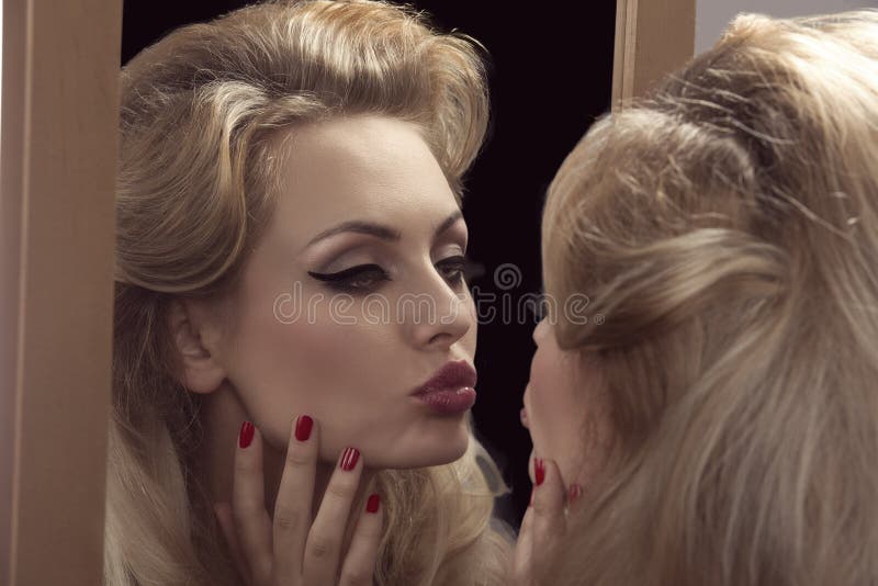 Close-up portrait of elegant blonde girl looking her cute make-up in to the mirror, posing with classic hair-style and red nail. Close-up portrait of elegant blonde girl looking her cute make-up in to the mirror, posing with classic hair-style and red nail