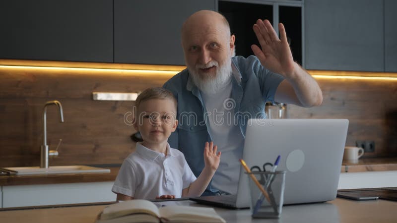 Niedlich und lustig Großvater und Enkel sitzen am Tisch mit Laptop Blick auf Kamera Lächeln und Welle an Betreiber. Hand
