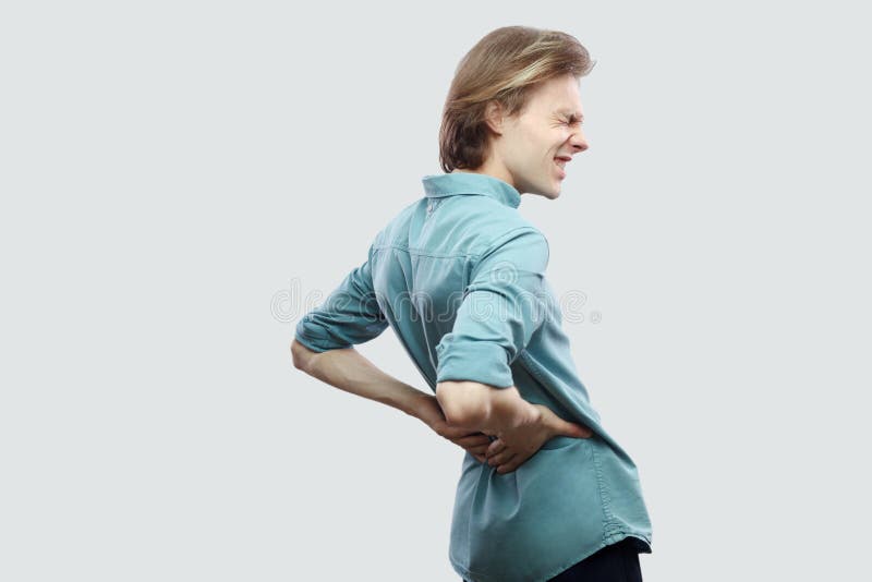 Kidney or spine pain. Profile side view portrait of handsome long haired blonde young man in blue casual shirt standing touching his painful back. indoor studio shot, isolated on light grey background. Kidney or spine pain. Profile side view portrait of handsome long haired blonde young man in blue casual shirt standing touching his painful back. indoor studio shot, isolated on light grey background