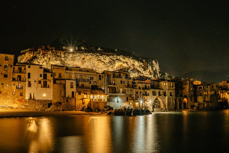 Night view of beach in Cefalu, Sicily, Italy, illuminated old town with colorful waterfront houses, sea and La Rocca cliff.