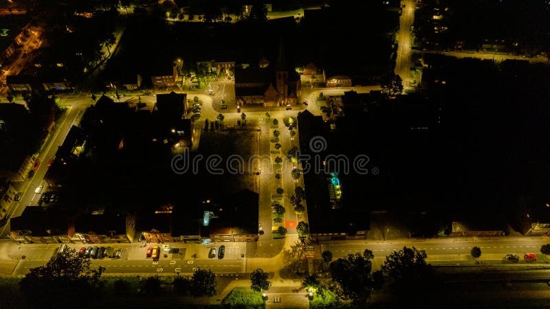 Această imagine antenă nocturnă capturează iluminarea ambiantă a unui pătrat orășenesc unde strălucirea călduroasă a luminilor stradale creează o rețea de vizibilitate pe fundalul întunecat al nopții. piața centrală, eventual ancorată de o biserică sau de o clădire semnificativă, devine un punct focal cu împrejurimile sale scăldate în lumină artificială. farurile vehiculelor și stopurile se strecoară pe străzi, urmărind traseele activității din seara trecută. siluetele întunecate ale copacilor și arhitectura înconjurătoare reprezintă scena care dă un sentiment de viață liniștită după amurg într-un mediu urban. această fotografie transmite caracterul pașnic, dar încă în viață al unui oraș în repaus. scenă urbană nocturnă : iluminarea pătrată a orașului de deasupra. fotografie de calitate superioară