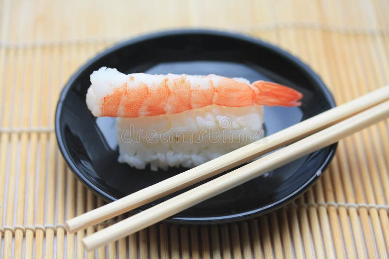 A traditional Nigirisushi, made from cooked vinegared sushi rice and a scrimp topping. This sushi is served on a small black plate and eaten with chopsticks. A traditional Nigirisushi, made from cooked vinegared sushi rice and a scrimp topping. This sushi is served on a small black plate and eaten with chopsticks