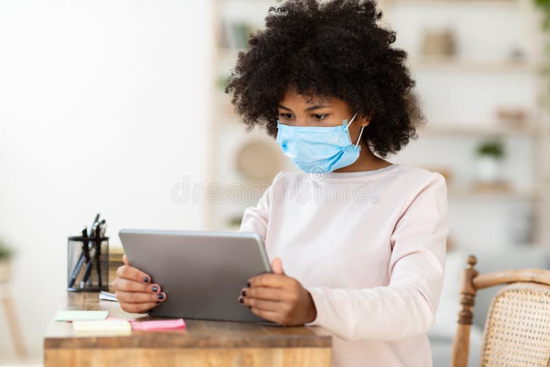 Niña adolescente africana que usa una tableta que usa máscara médica en casa foto de archivo