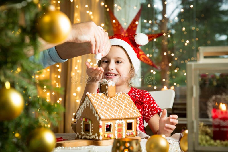 Niña adorable en sombrero rojo que adorna la casa de pan de jengibre de la Navidad con el esmalte foto de archivo libre de regalías Niña adorable en sombrero rojo que adorna la casa de pan de jengibre de la Navidad con el esmalte foto de archivo libre de regalías