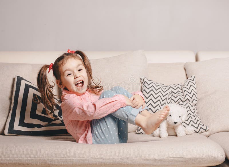 Niña bastante feliz en sentarse que lleva casual en el sofá con el perro de juguete y la sonrisa imagenes de archivo Niña bastante feliz en sentarse que lleva casual en el sofá con el perro de juguete y la sonrisa imagenes de archivo