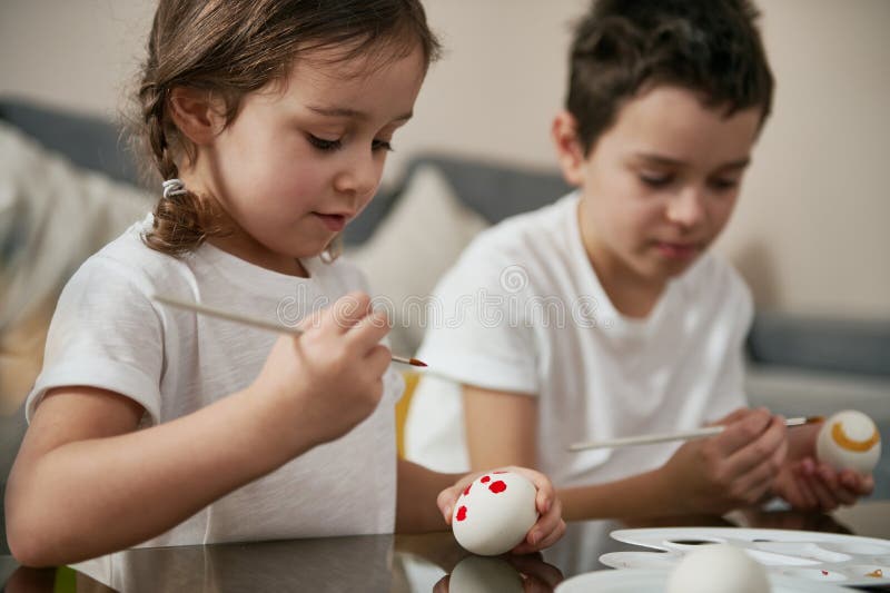 Niña de preescolar sentada al lado de su hermano usa pincel para decorar huevos de pascua. listos para celebrar la tradicional fotografía de archivo