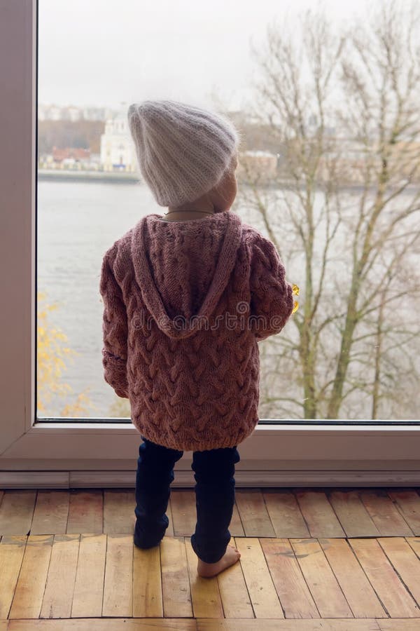 Little girl in a purple sweater and cap standing at the window with his back. Little girl in a purple sweater and cap standing at the window with his back