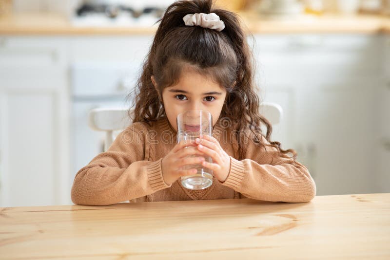 Niña linda beber agua de vidrio sentado en la mesa en la cocina fotografía de archivo libre de regalías