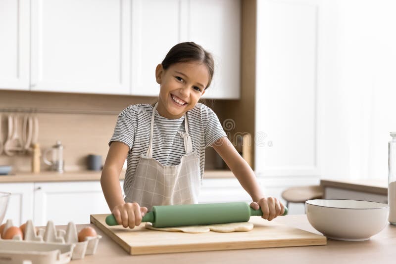 Happy beautiful baker kid girl in household apron preparing bakery meal in kitchen, cooking pastry dessert for family dinner, rolling raw dough on table, looking at camera, smiling for portrait. Happy beautiful baker kid girl in household apron preparing bakery meal in kitchen, cooking pastry dessert for family dinner, rolling raw dough on table, looking at camera, smiling for portrait