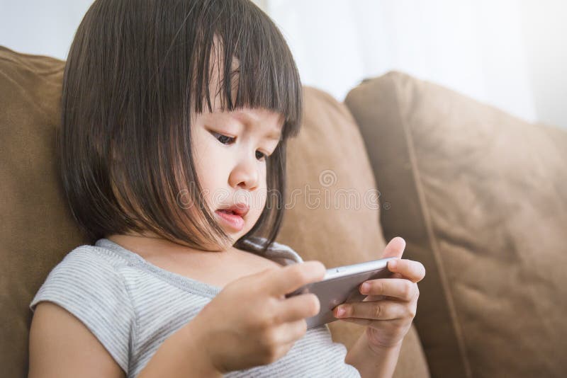 Niña pequeña asiática casual que se sienta en un sofá en casa que juega y que toca un teléfono móvil imagen de archivo libre de regalías Niña pequeña asiática casual que se sienta en un sofá en casa que juega y que toca un teléfono móvil imagen de archivo libre de regalías