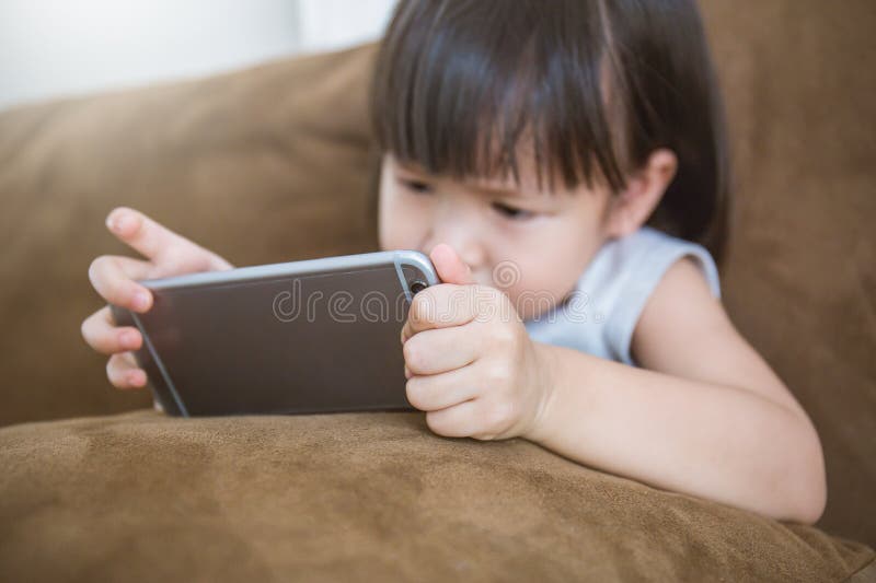 Niña pequeña asiática casual que se sienta en un sofá en casa que juega y que toca un teléfono móvil fotos de archivo Niña pequeña asiática casual que se sienta en un sofá en casa que juega y que toca un teléfono móvil fotos de archivo