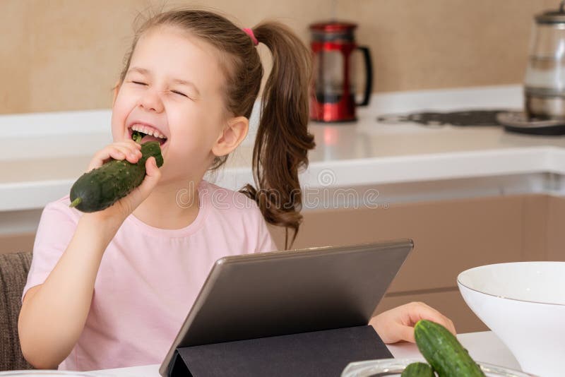 Niña Pequeña Comiendo Pepino Y Usando El Tablet En La Cocina Foto De Archivo Imagen De 