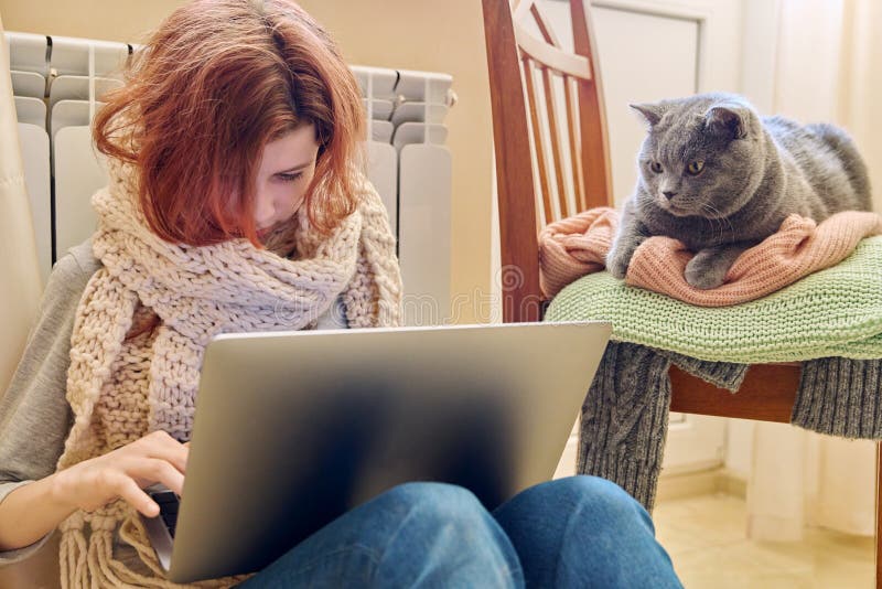Niña preadolescente con pañuelo usando computadora portátil calentando cerca de radiador de calefacción con gato gris mascota foto de archivo