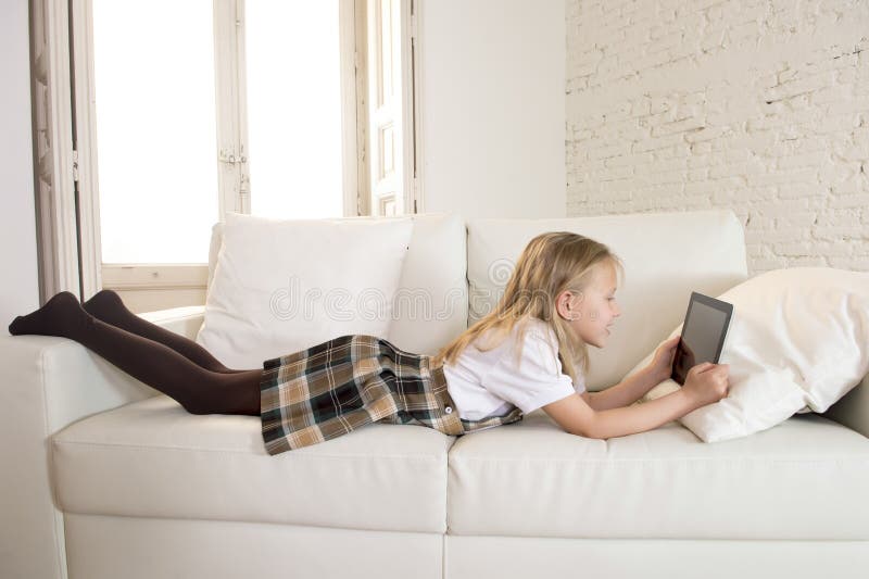 Niña rubia que miente en el sofá casero del sofá usando Internet app en el cojín digital de la tableta en el cojín digital de la imagen de archivo