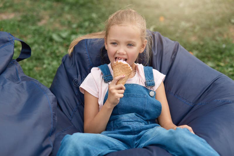Niña rubia sentada en una silla azul sin marco en el parque y comiendo helado Feliz bebé comiendo helado mientras se relaja fotos de archivo libres de regalías Niña rubia sentada en una silla azul sin marco en el parque y comiendo helado Feliz bebé comiendo helado mientras se relaja fotos de archivo libres de regalías