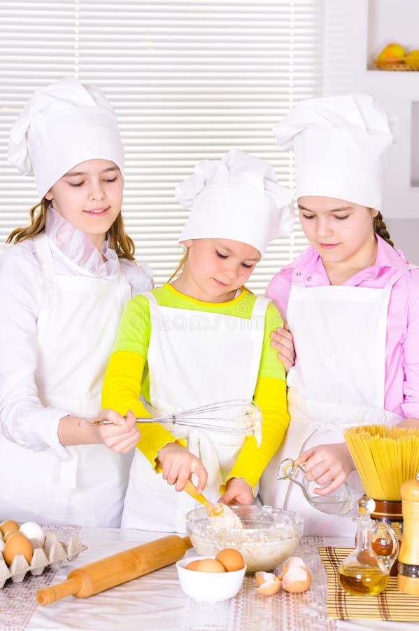 Niñas lindas en sombreros del ` s del cocinero y delantales que preparan la pasta en la cocina imagen de archivo Niñas lindas en sombreros del ` s del cocinero y delantales que preparan la pasta en la cocina imagen de archivo