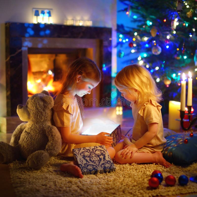 Adorable little girls opening a magical Christmas gift by a Christmas tree in cozy living room in winter. Adorable little girls opening a magical Christmas gift by a Christmas tree in cozy living room in winter