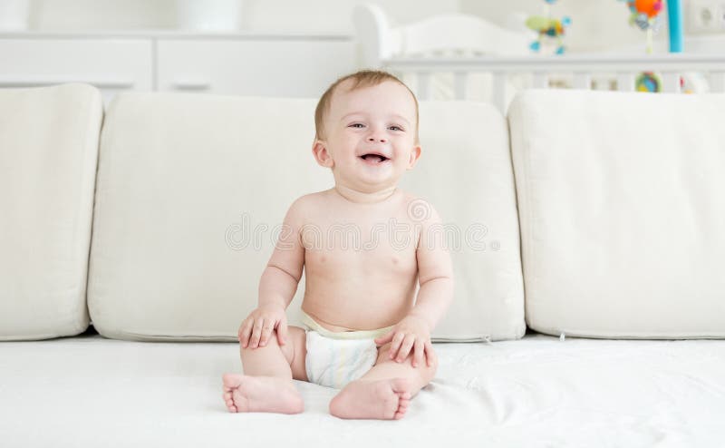 Niño pequeño de risa feliz en los pañales que se sientan en el sofá y que miran in camera fotos de archivo libres de regalías Niño pequeño de risa feliz en los pañales que se sientan en el sofá y que miran in camera fotos de archivo libres de regalías