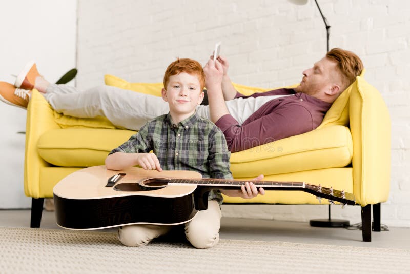 Niño pequeño lindo que sostiene el anuncio de la guitarra acústica que sonríe en la cámara mientras que padre usando la tableta d foto de archivo