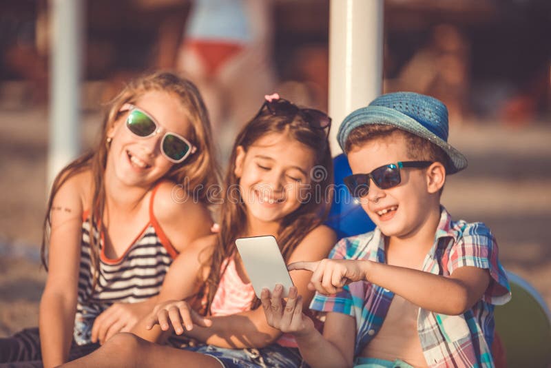 Niño pequeño lindo y muchachas que se sientan en la playa que juega con el teléfono elegante imagen de archivo Niño pequeño lindo y muchachas que se sientan en la playa que juega con el teléfono elegante imagen de archivo