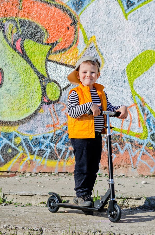 Niño pequeño orgulloso con su vespa que se coloca en sol con su gorra de béisbol al ángulo libertino que sonríe en la cámara, contexto de la pared cubierto en pintada colorida