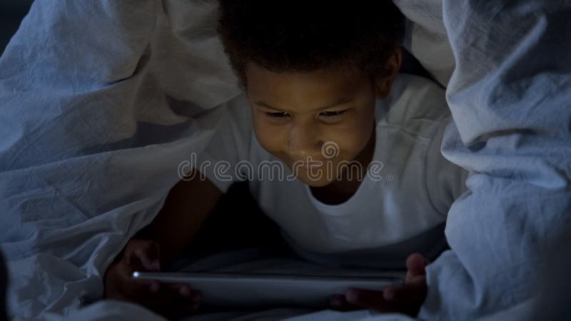 Niño que mira la tableta secretamente la noche, cubierta con la cabeza en manta foto de archivo Niño que mira la tableta secretamente la noche, cubierta con la cabeza en manta foto de archivo
