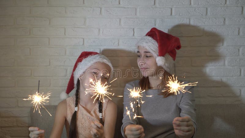 Niños alegres, felices en los sombreros de Papá Noel sostener bengalas encendidas en sus manos, agitarlas y sonreír sentándose en almacen de video