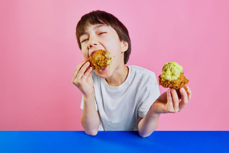 Moment of pleasure. Little boy, child in white t-shirt sitting at table and emotionally eating fried chicken with mustard sauce against pink background. Concept of food, childhood, emotions, pop art. Moment of pleasure. Little boy, child in white t-shirt sitting at table and emotionally eating fried chicken with mustard sauce against pink background. Concept of food, childhood, emotions, pop art