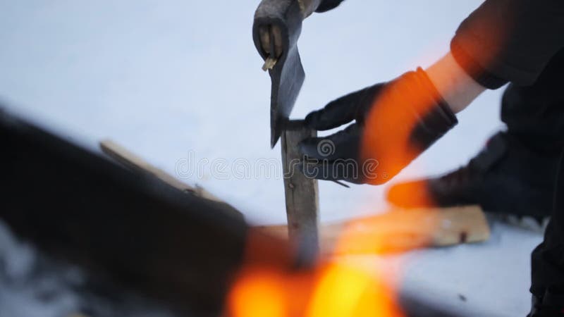 No inverno, luvas vestindo de um homem que desbastam a madeira com um machado em um fundo do fogo