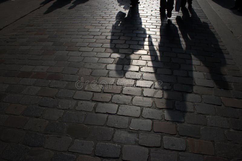 A picture of a cobblestone alley, dimly lit in the night with the feet and shadows of a few pedestrians visible. A picture of a cobblestone alley, dimly lit in the night with the feet and shadows of a few pedestrians visible.