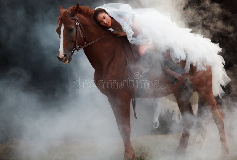 Noiva bonita da beleza na equitação nupcial branca do traje do casamento da forma no cavalo muscular forte no fundo sonhador pret fotos de stock Noiva bonita da beleza na equitação nupcial branca do traje do casamento da forma no cavalo muscular forte no fundo sonhador pret fotos de stock