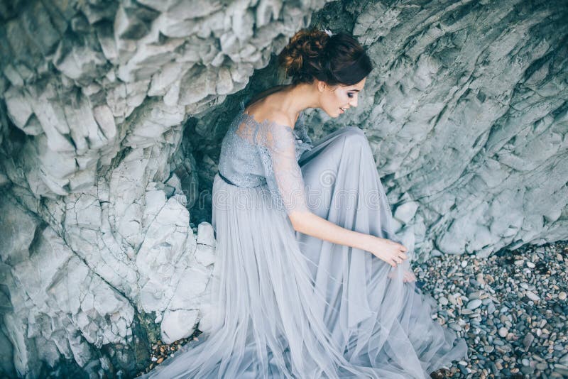 A noiva bonita da menina em um vestido cinzento que senta-se pelo mar perto da rocha sorri, ri, endireita seu vestido do laço imagens de stock royalty free A noiva bonita da menina em um vestido cinzento que senta-se pelo mar perto da rocha sorri, ri, endireita seu vestido do laço imagens de stock royalty free