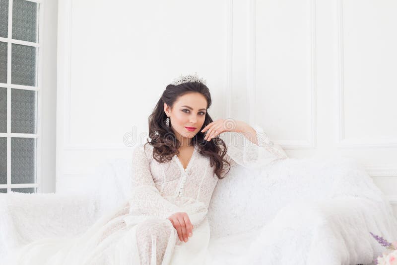 A noiva em um vestido de casamento com uma coroa que senta-se em um sofá branco imagens de stock A noiva em um vestido de casamento com uma coroa que senta-se em um sofá branco imagens de stock