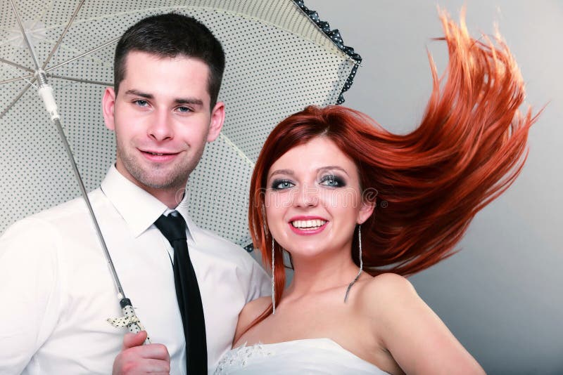 Wedding day. Portrait of happy married couple blue eyed bride with her red hair blowing in the wind and groom with umbrella studio shot on gray background. Wedding day. Portrait of happy married couple blue eyed bride with her red hair blowing in the wind and groom with umbrella studio shot on gray background