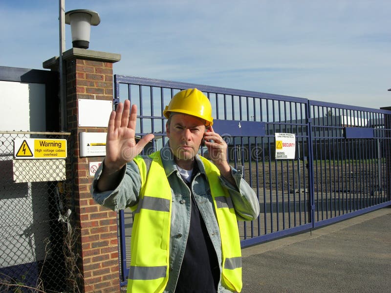A work site gate supervisor wearing hige visability vest and yellow hard hat raises his hand to stop visitors while using his mobile phone to get more instructions. A work site gate supervisor wearing hige visability vest and yellow hard hat raises his hand to stop visitors while using his mobile phone to get more instructions.