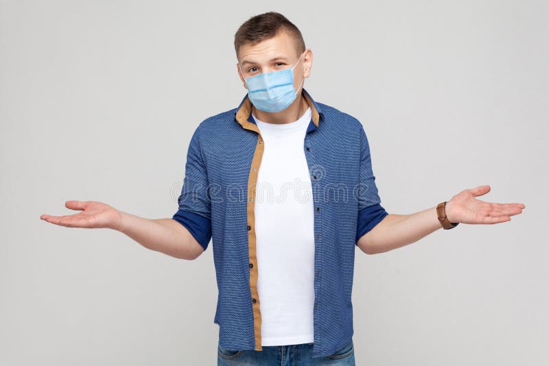I don`t know. Portrait of thoughtful young man in casual style with surgical medical mask standing, raised arms and looking at camera thinking. indoor studio shot, isolated on gray background. I don`t know. Portrait of thoughtful young man in casual style with surgical medical mask standing, raised arms and looking at camera thinking. indoor studio shot, isolated on gray background