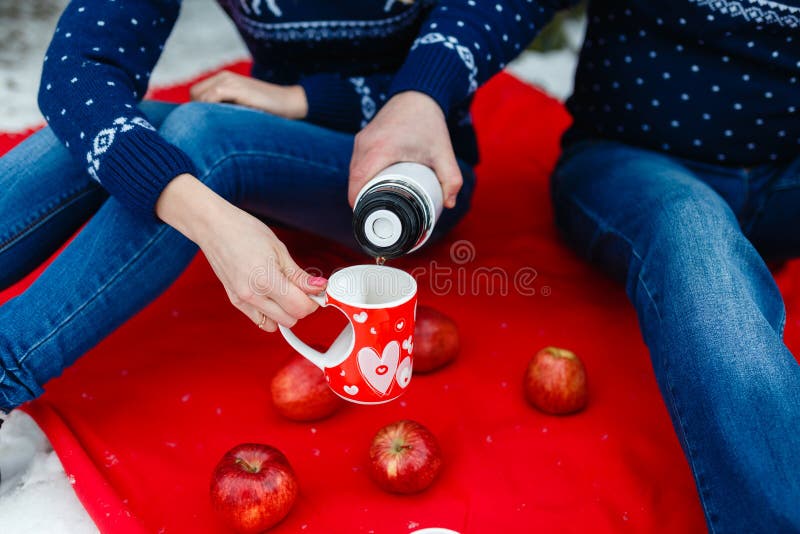 Το άτομο χύνει το τσάι σε μια κούπα με μια εικόνα της καρδιάς Τα νέα ζεύγη στα πουλόβερ κάθονται στο κόκκινο κάλυμμα Γαμήλιο πικ- στοκ εικόνες με δικαίωμα ελεύθερης χρήσης