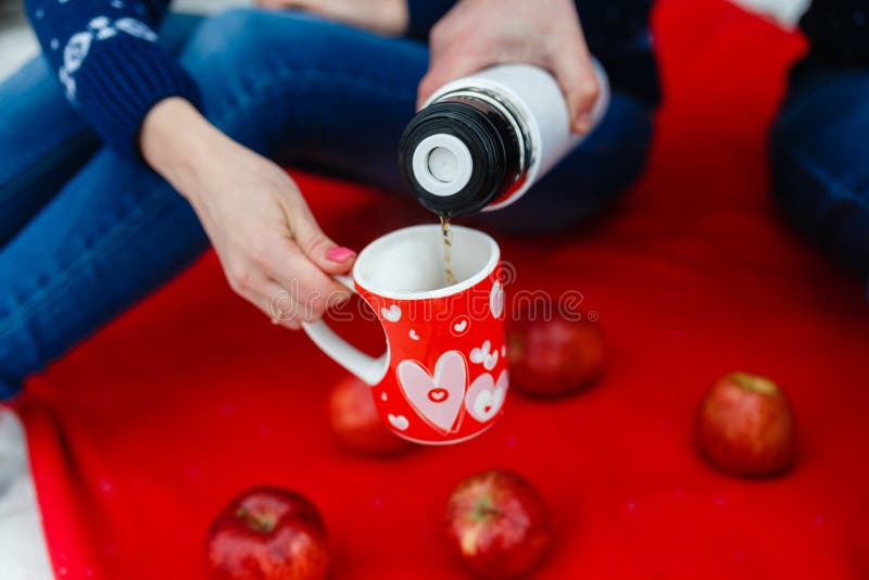 Το άτομο χύνει το τσάι σε μια κούπα με μια εικόνα της καρδιάς Τα νέα ζεύγη στα πουλόβερ κάθονται στο κόκκινο κάλυμμα Γαμήλιο πικ- στοκ φωτογραφία με δικαίωμα ελεύθερης χρήσης