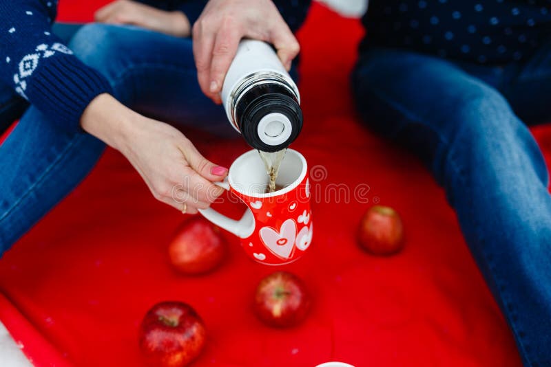 Το άτομο χύνει το τσάι σε μια κούπα με μια εικόνα της καρδιάς Τα νέα ζεύγη στα πουλόβερ κάθονται στο κόκκινο κάλυμμα Γαμήλιο πικ- στοκ φωτογραφίες με δικαίωμα ελεύθερης χρήσης