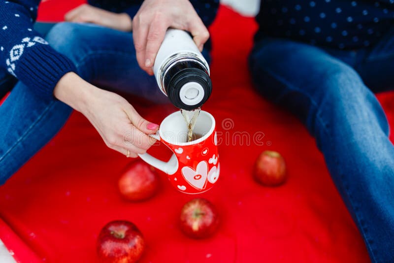 Το άτομο χύνει το τσάι σε μια κούπα με μια εικόνα της καρδιάς Τα νέα ζεύγη στα πουλόβερ κάθονται στο κόκκινο κάλυμμα Γαμήλιο πικ- στοκ φωτογραφία με δικαίωμα ελεύθερης χρήσης