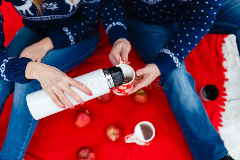 Το κορίτσι χύνει το τσάι σε μια κούπα με μια εικόνα της καρδιάς Τα νέα ζεύγη στα πουλόβερ κάθονται στο κόκκινο κάλυμμα Γαμήλιο πι στοκ φωτογραφία με δικαίωμα ελεύθερης χρήσης
