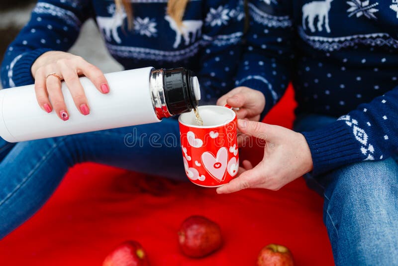Το κορίτσι χύνει το τσάι σε μια κούπα με μια εικόνα της καρδιάς Τα νέα ζεύγη στα πουλόβερ κάθονται στο κόκκινο κάλυμμα Γαμήλιο πι στοκ φωτογραφία