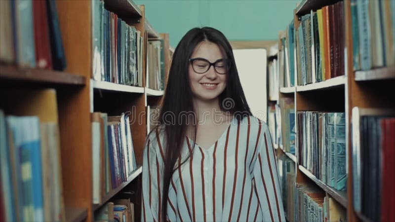 Το όμορφο νέο κορίτσι με τη μακριά μαύρη τρίχα πηγαίνει μεταξύ των κάθετων ραφιών στη βιβλιοθήκη και χαμογελά φιλμ μικρού μήκους
