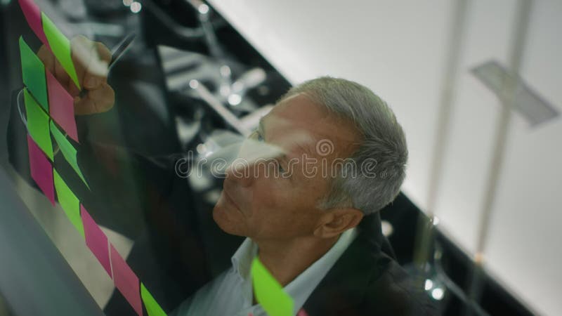 ένας επιχειρηματίας γράφει αυτοκόλλητα σημειώματα σε γυάλινη σανίδα στο γραφείο απόθεμα βίντεο