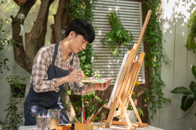 一位年轻快乐的亚洲男性艺术家穿着围裙，在他的家庭花园里在一张画架上的画布上着色。 库存照片