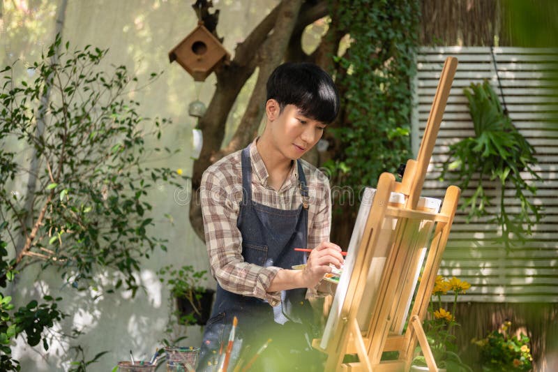 年轻快乐的亚洲男性艺术家穿着围裙在自家花园里的画架上着色画布 免版税库存图片